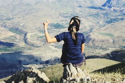 Rear view of woman showing peace sign while sitting on cliff