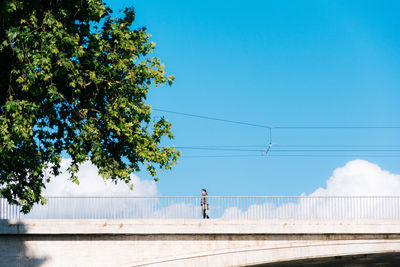 Low angle view of woman walking on bridge by tree against blue sky