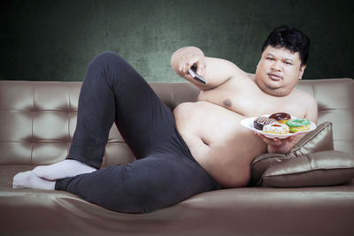 Portrait of shirtless man holding donuts in plate while leaning on sofa
