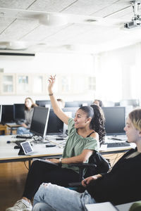 Smiling teenage girl sitting with hand raised by young friend in computer lab at high school