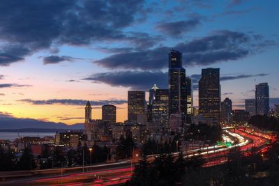 Illuminated highway amidst buildings against sky during sunset