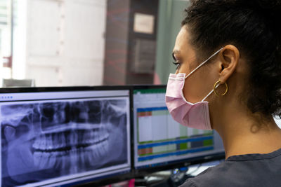 Side view of black female doctor in medical mask examining x ray on monitor of computer while working in modern dental clinic