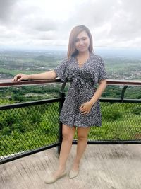 Portrait of young woman standing on railing against sky