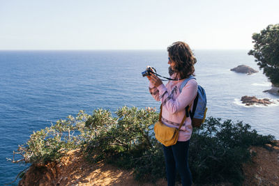 Side view of young woman photographing while standing on mountain against sea