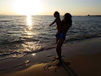 Silhouette mother carrying son while standing at beach during sunset against sky