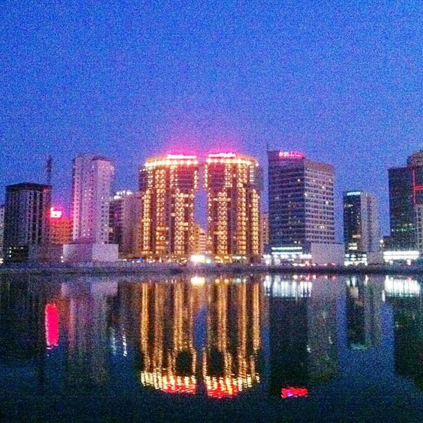 water, building exterior, reflection, waterfront, architecture, city, built structure, illuminated, skyscraper, cityscape, sky, blue, modern, urban skyline, night, river, clear sky, office building, multi colored, sea