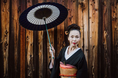 Portrait of woman holding umbrella standing against wooden wall