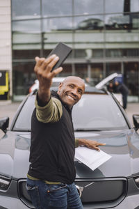 Side view of mature man taking selfie through mobile phone with new car