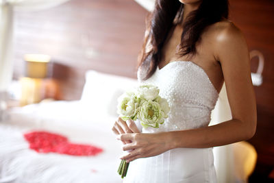 Midsection of bride holding white flower while standing in bedroom at home