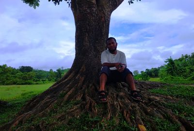 Full length of man sitting by tree against sky