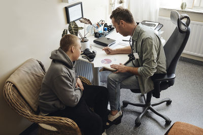 High angle view of school male discussing over chart with nurse student sitting at table in office