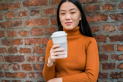 Portrait of beautiful young woman holding disposable cup against brick wall
