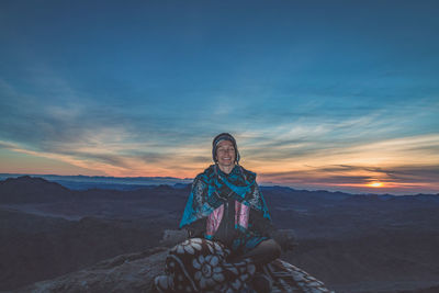 Woman meditating while sitting on rock against sky during sunset