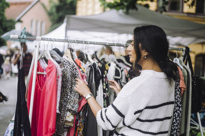 Young woman choosing dress hanging on clothes rack at flea market