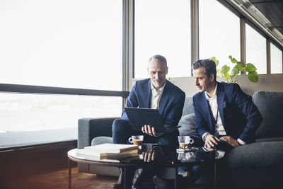 Businessman showing laptop to colleague while sitting on sofa at workplace