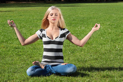 Full length of young woman meditating while sitting on grassy field in park