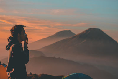 Side view of man smoking by mountains during sunrise