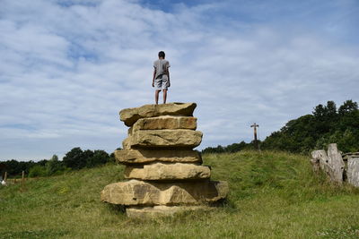 Rear view of boy standing on rocks over field against cloudy sky