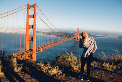 Woman standing by suspension bridge against sky during sunset