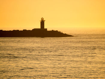 Silhouette lighthouse by sea against orange sky