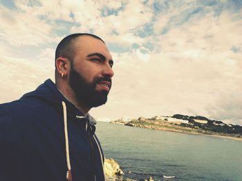 Young man looking away while standing by sea against cloudy sky