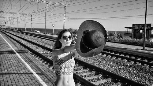 Young woman wearing sunglasses holding hat while standing on railway station against sky