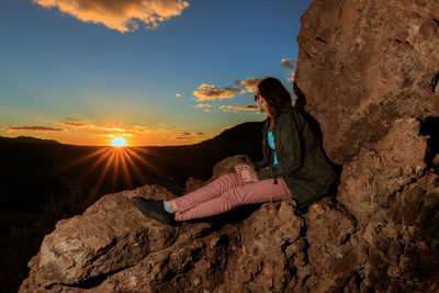 Side view full length of woman sitting on rock formation against sky during sunset