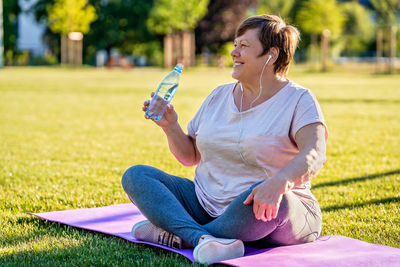 Senior plus size woman sitting on yoga mat on green grass outdoors resting  drinking water 