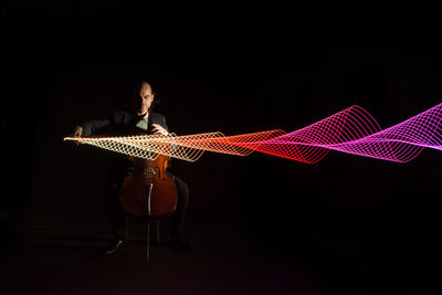 Full length of musician playing cello by light painting against black background