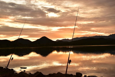 Silhouette fishing rod by lake against sky during sunset