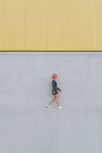 Side view of young female with bright pink hair in stylish outfit walking on border at gray wall on street