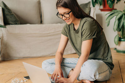 Smiling european female student in glasses communicates on a laptop while sitting on the floor.