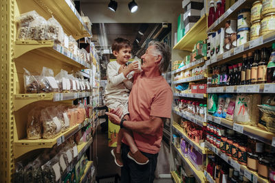 Smiling senior man carrying grandson while shopping snacks in supermarket