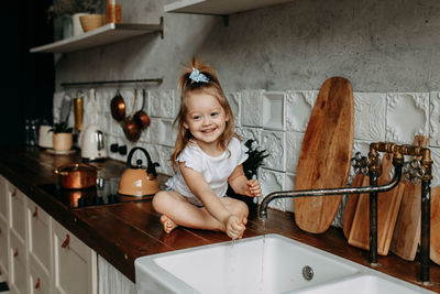 A cheerful little girl child sits on the kitchen table and plays splashing water from the sink