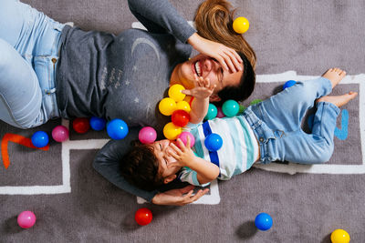 From above delighted woman and little boy screaming and throwing colorful balls up while lying on nursery floor together