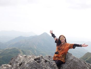 Portrait of smiling young woman with arms raised on mountain against sky