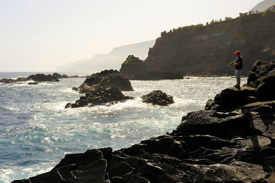 Man standing on rock by sea against sky