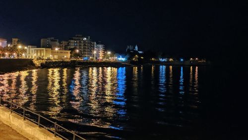 Illuminated buildings by sea against sky at night