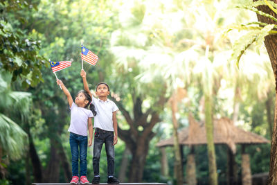 Siblings holding malaysian flag while standing on bench against trees at park