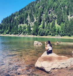 Girl sitting on a rock taking a picture, on mountain and lake background 