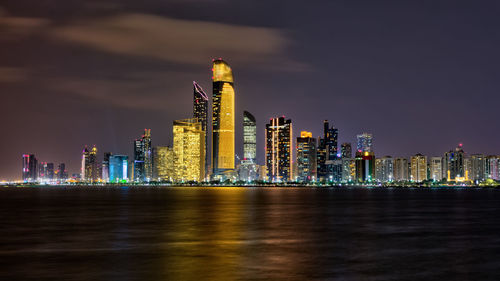 Illuminated modern buildings by sea against sky at night