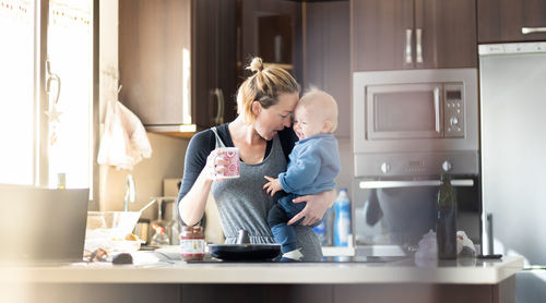 Happy mother and little infant baby boy together making pancakes for breakfast in domestic kitchen