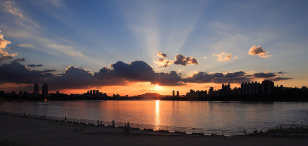 Silhouette buildings by river against sky during sunset