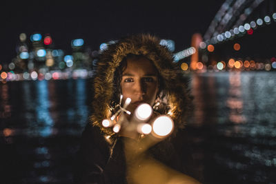 Portrait of young woman holding illuminated light while standing against river at night