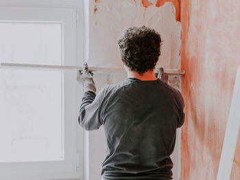 A young caucasian guy smoothes fresh putty on the wall near the window with a wooden rule