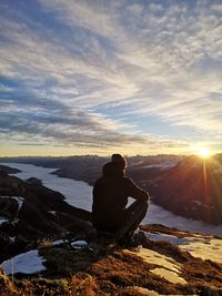 Rear view of man sitting on rock against sky during sunset