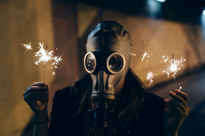 Woman holding illuminated sparklers while wearing gas mask 