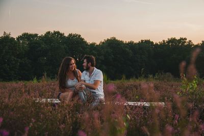 Young couple sitting in field against sky during sunset