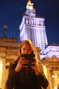 Low angle view of woman using smart phone against illuminated palace of culture and science at night