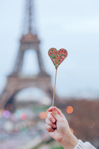 Cropped hand of woman holding heart shape candy against eiffel tower